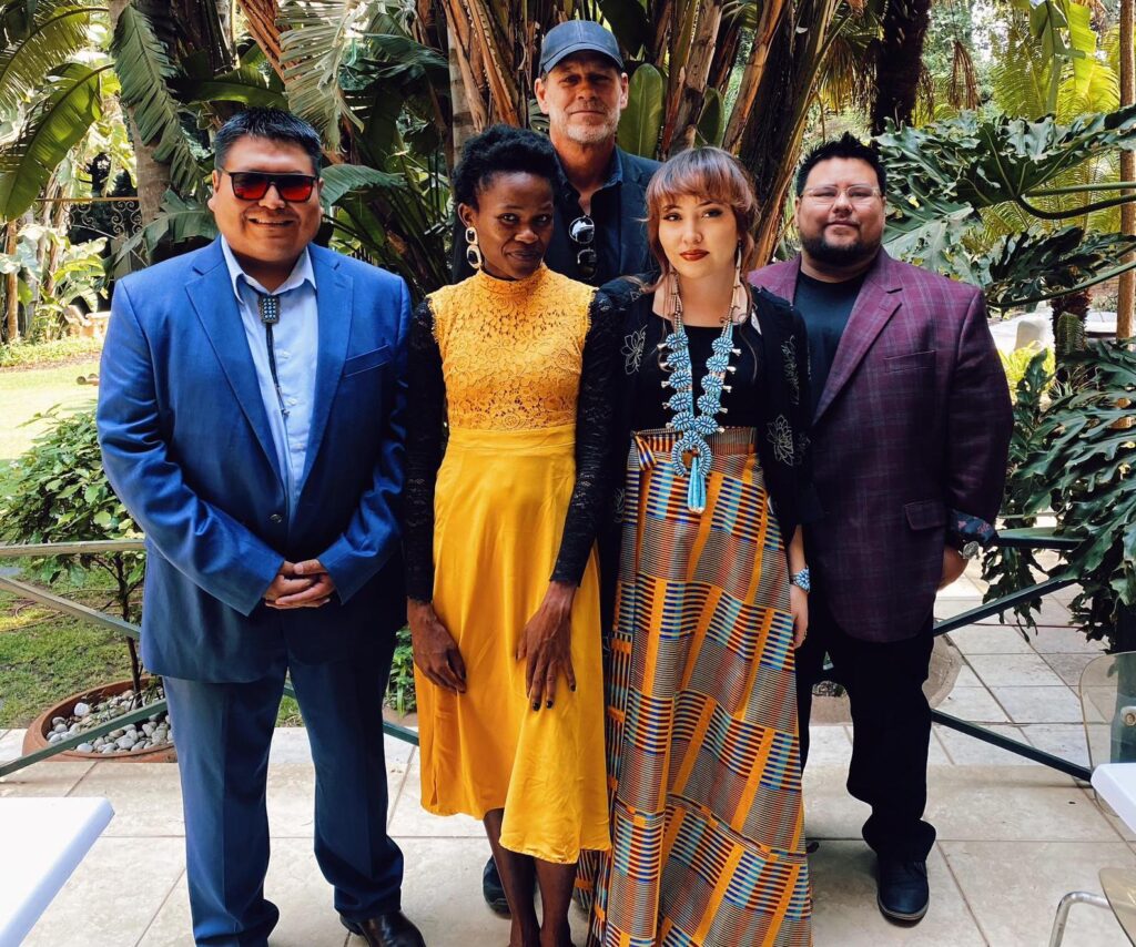 A photograph features five people in colorful clothing standing on cement tiles in front of tall trees and green grass.
