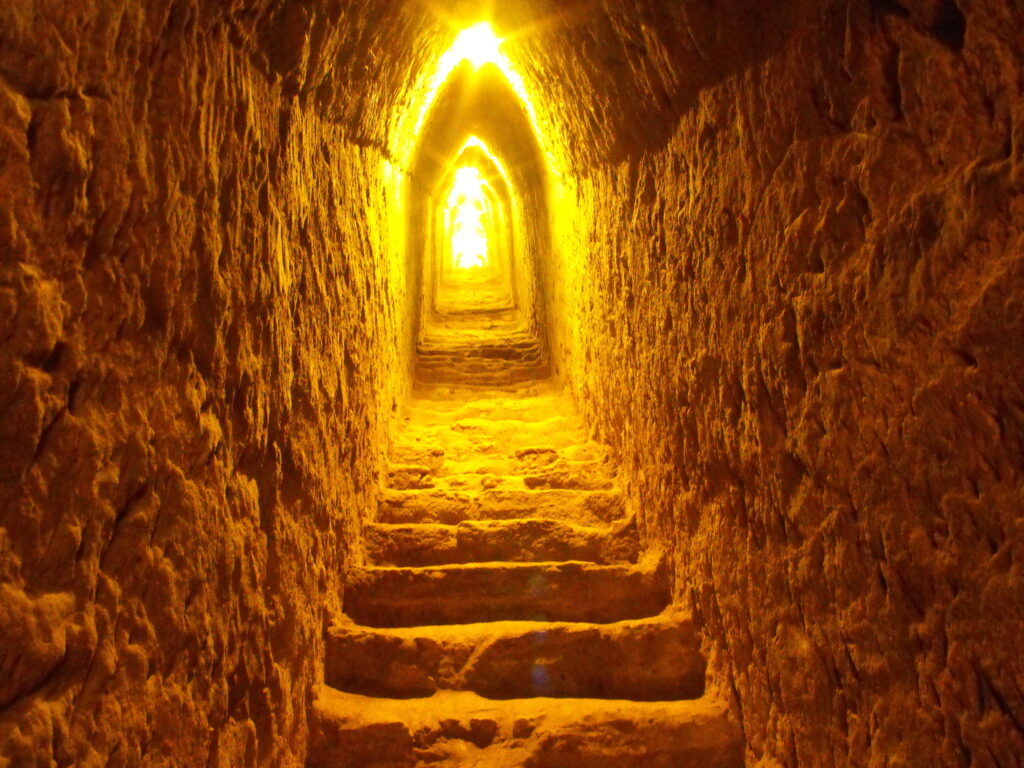 A photograph features a brown stone staircase lined with rough walls of similar stone. A bright yellow light illuminates the far side of a tunnel-like perspective toward the staircase’s top end.