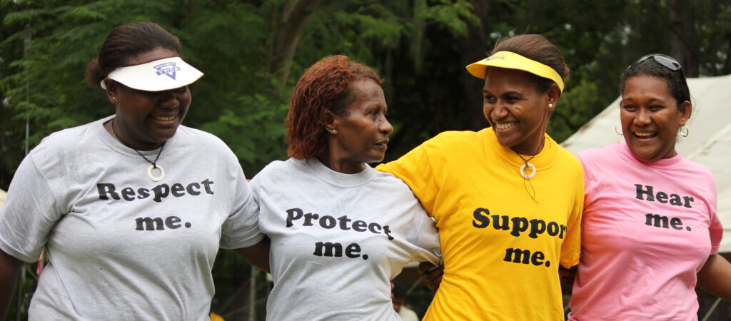 A photograph features four women standing with their arms over the shoulders of the women to their immediate left and/or right. From left to right, the women wear shirts that respectively read, in black text on white, yellow, or pink shirts: “Respect me.” “Protect me.” “Support me.” and “Hear me.”
