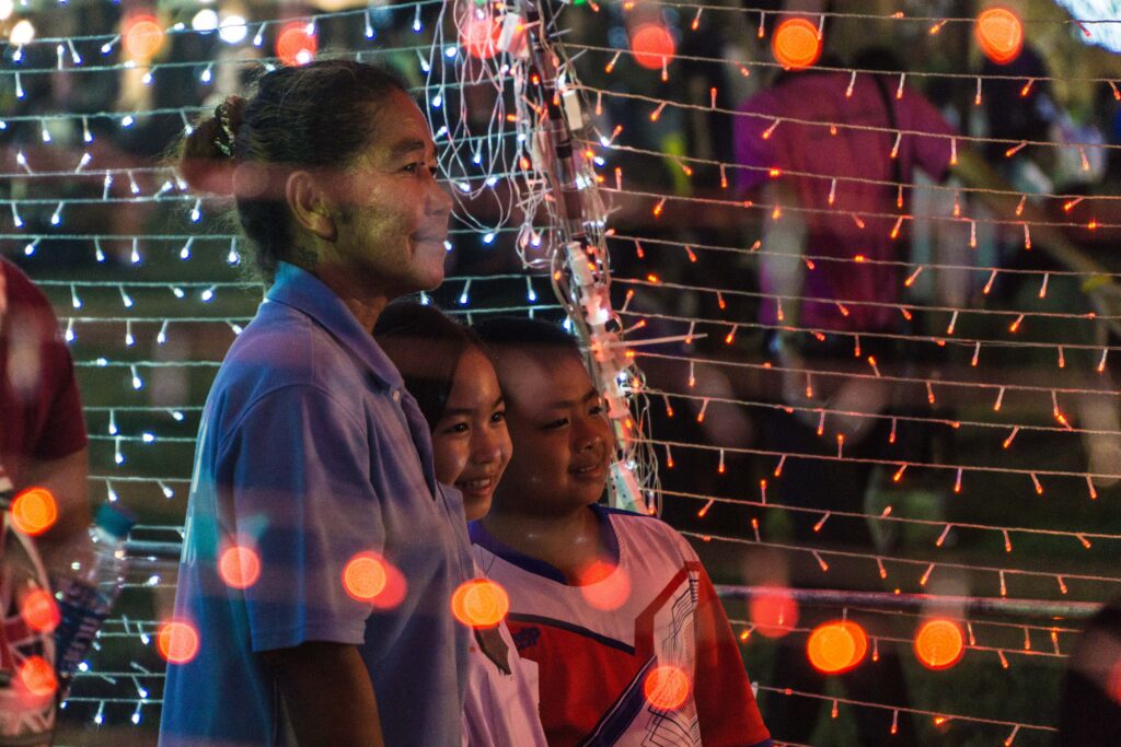 A smiling older woman stands close to two smiling children surrounded by strings of small orange lights hung in rows.