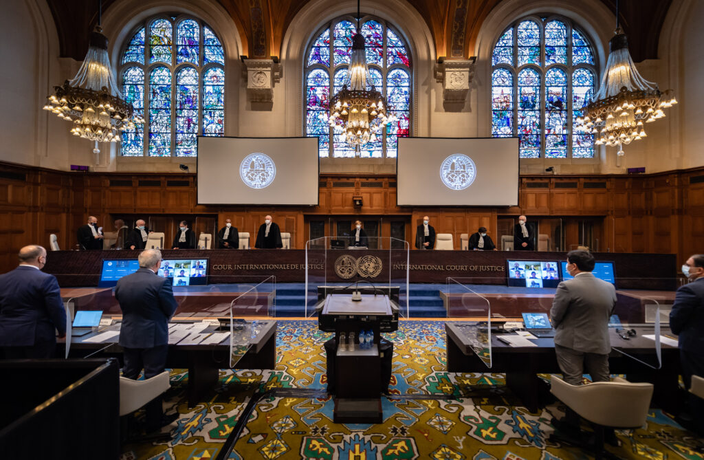 A courtroom features a lectern surrounded by glass and people standing behind a long wooden desk below stained glass.