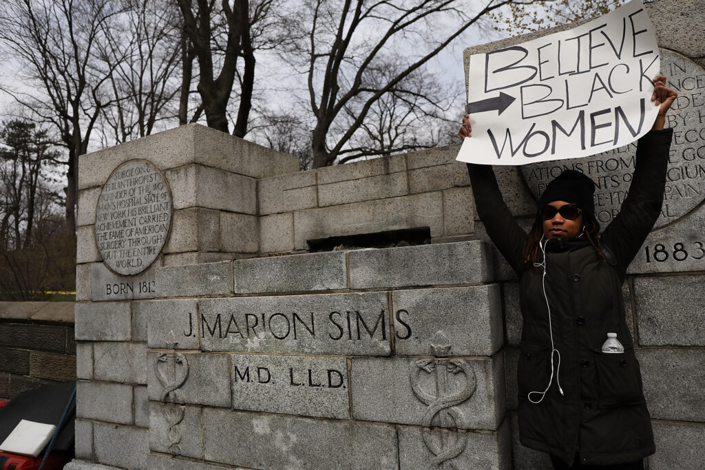 A photograph features a woman wearing a black jacket and sunglasses standing next to an empty stone pedestal with text carved into it that reads, "J. Marion Sims M.D., L.L.D." The woman holds a sign that reads, "BELIEVE BLACK WOMEN."