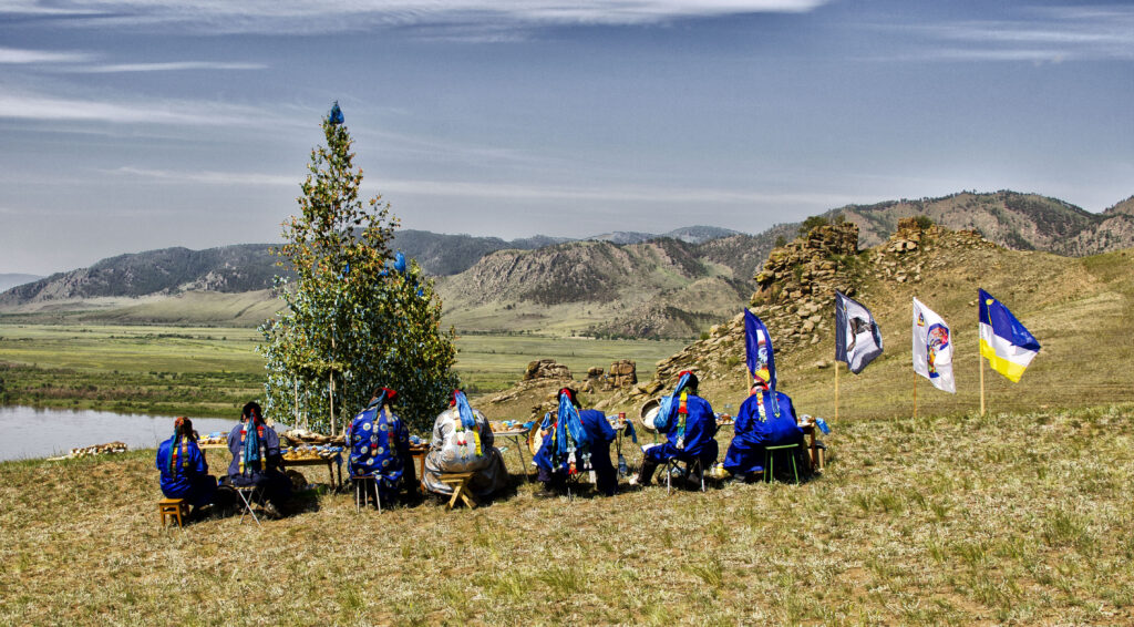 A photograph features seven people seated on a grassy plain with a tree and body of water to their left, four flags posted on their right, and mountains and blue skies in the distance. All are wearing headpieces and blue clothing decorated with colorful patches.