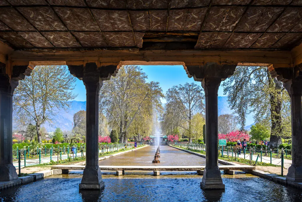 A photograph taken from under a large building’s awning looks out to show a fountain with green and red tress lining either side. The building’s stone pillars line the foreground.