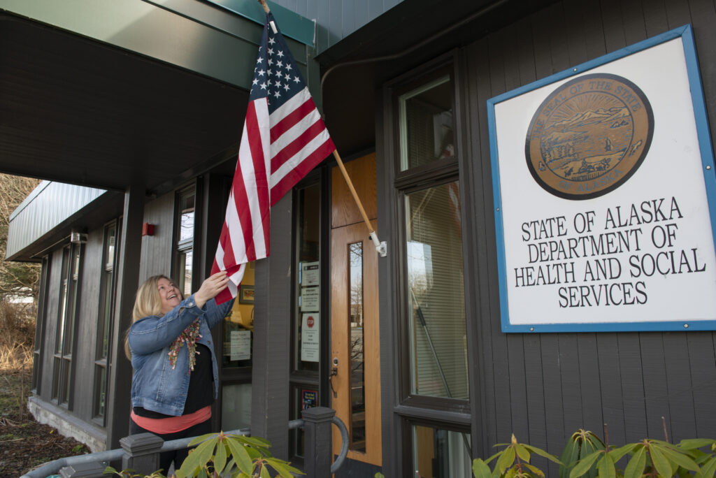 A photograph features the front of a gray building with an American flag hung on the wall and a large sign that reads, “STATE OF ALASKA DEPARTMENT OF HEALTH AND SOCIAL SERVICES.” A person with shoulder-length blonde hair wears denim jacket and reaches up to adjust the flag.