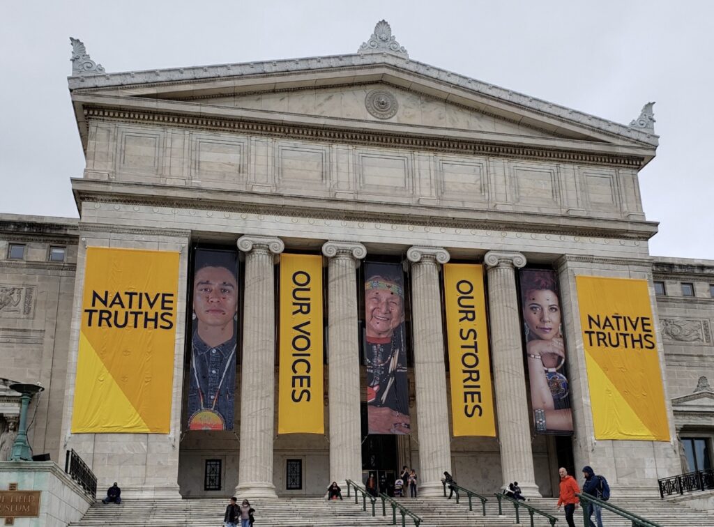 A photograph features a large building with four pillars across its front side. Between the pillars hang three long banners of people’s portraits alternately separated by yellow banners that read “Native Truths,” “Our Voices,” Our Stories” and “Native Truths.”