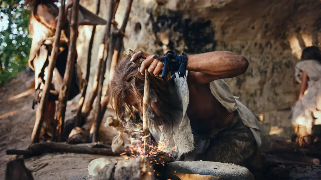 A photograph features a person wearing torn beige clothing kneeling in the front of a cave over a stone, rubbing wooden sticks together atop the latter to create sparks. Another brown-haired person in beige clothing is barely visible in the background on the right side of the image.