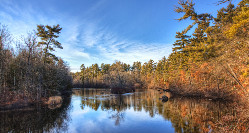 A photograph features a wide river lined on both sides with trees ranging in color from green to orange to yellow. Topped with blues skies, the trees along the back of the image are optically reflected in the water below.