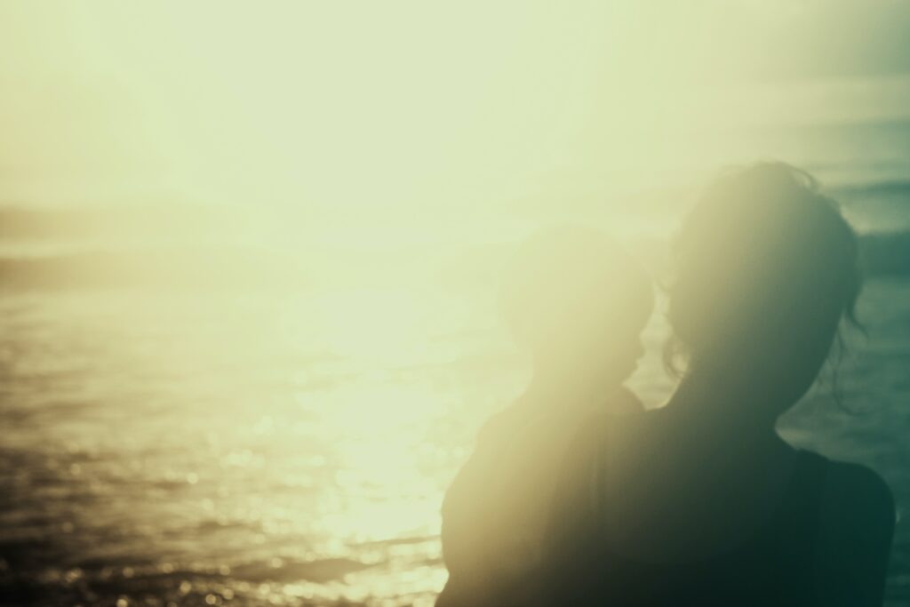 A photograph features a person with short, wavy hair holding a child near an ocean with a bright line shining from the background, blowing out an otherwise dark image.