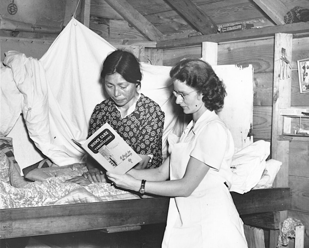 A black-and-white photograph features two people, one seated in a bunk bed and the other standing beside them. The standing person wears a white-collared dress and holds up a booklet for them both to look through.