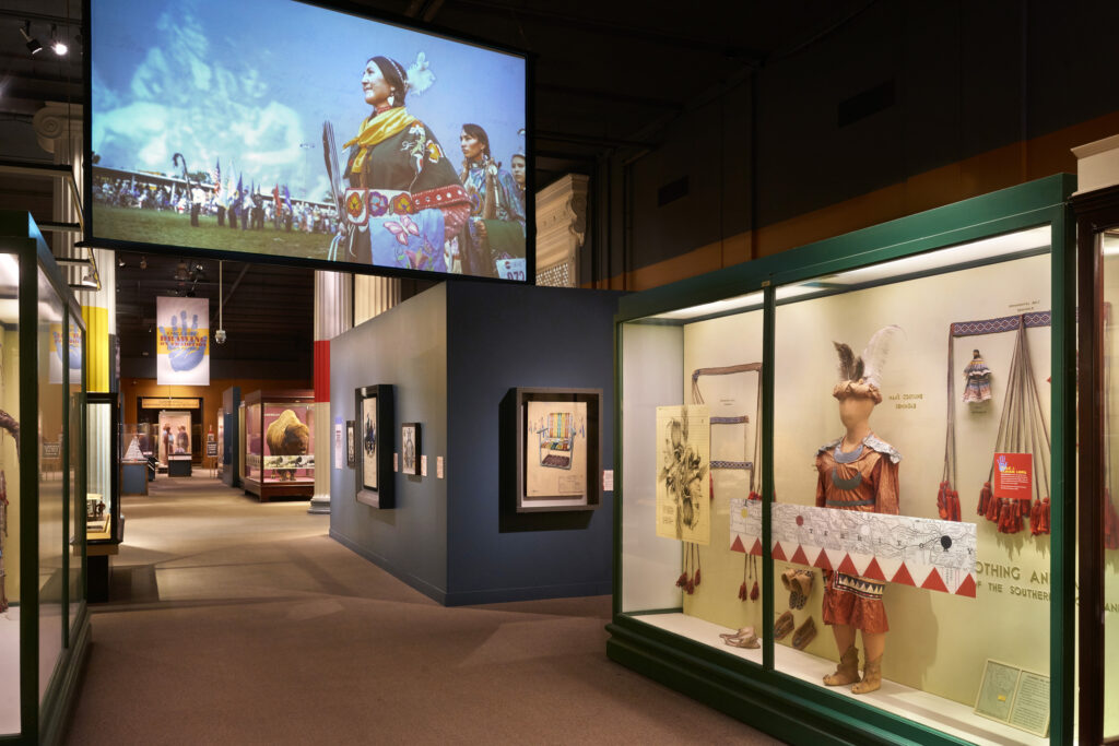 A photograph features a long hallway lined by museum cases that display Native American clothing such as headpieces, jewelry, garments, and shoes. Farther down the hallway, framed pictures hang on a dark blue wall and beyond that, a large case holds a stuffed buffalo.