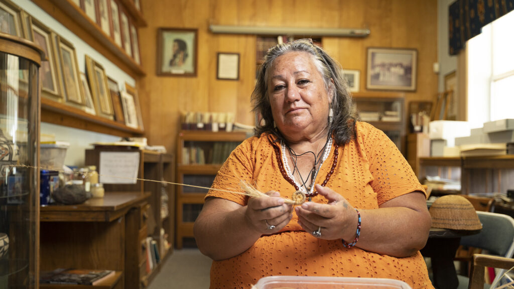 A photograph features a person with straight, gray, shoulder-length hair wearing an orange shirt and beaded jewelry. Holding a long thread connected to something out of frame taut, they smile and look straight ahead. An office setting and woven object are visible in the background.