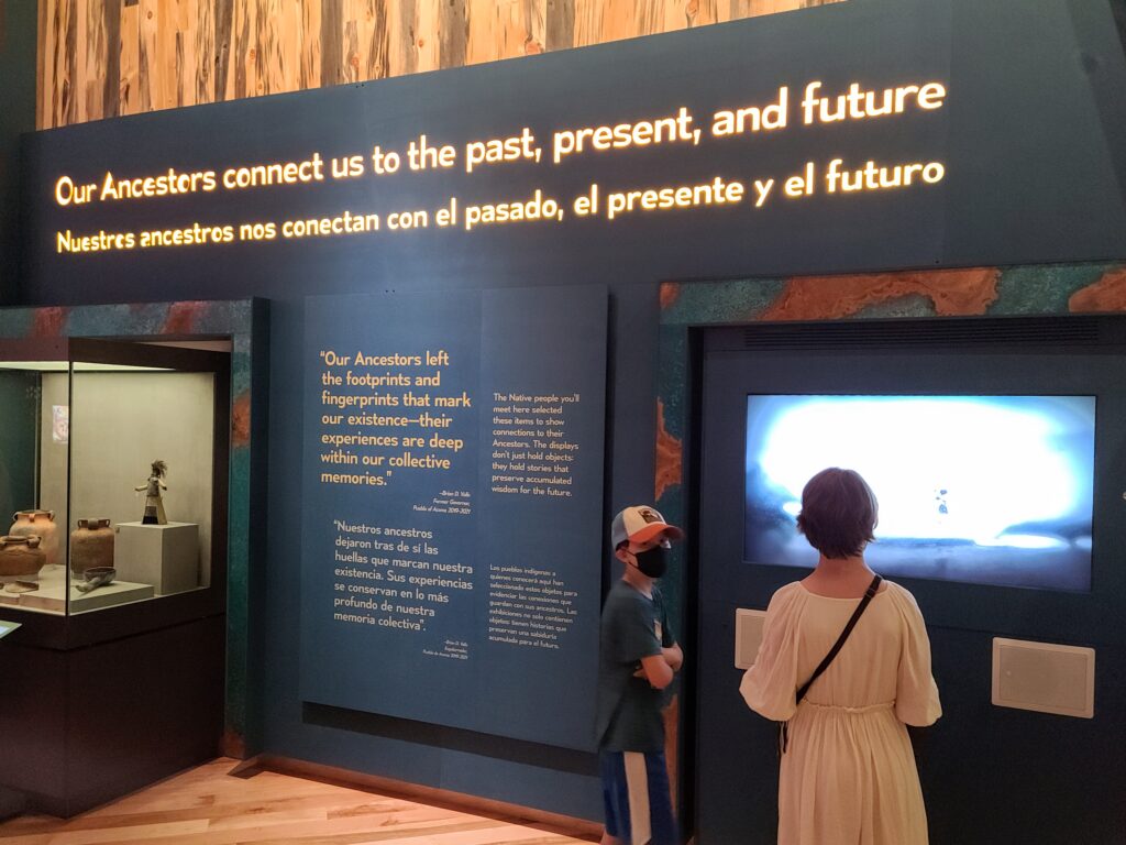 A photograph features wall text in English and Spanish between a museum case displaying pottery and an installed video screen that an adult and child are watching. Large white text at the top reads, “Our Ancestors connect us to the past, present, and future.” Underneath, a smaller quote reads, “’Our Ancestors left the footprints and fingerprints that mark our existence—their experiences are deep within our collective.’—Brian D. Vallo, Former Governor, Pueblo of Acoma 2019–2021.” To the right, a white text block reads, “The Native people you’ll meet here selected these items to show connections to their Ancestors. The displays don’t just hold objects: they hold stories that preserve accumulated wisdom for the future.”