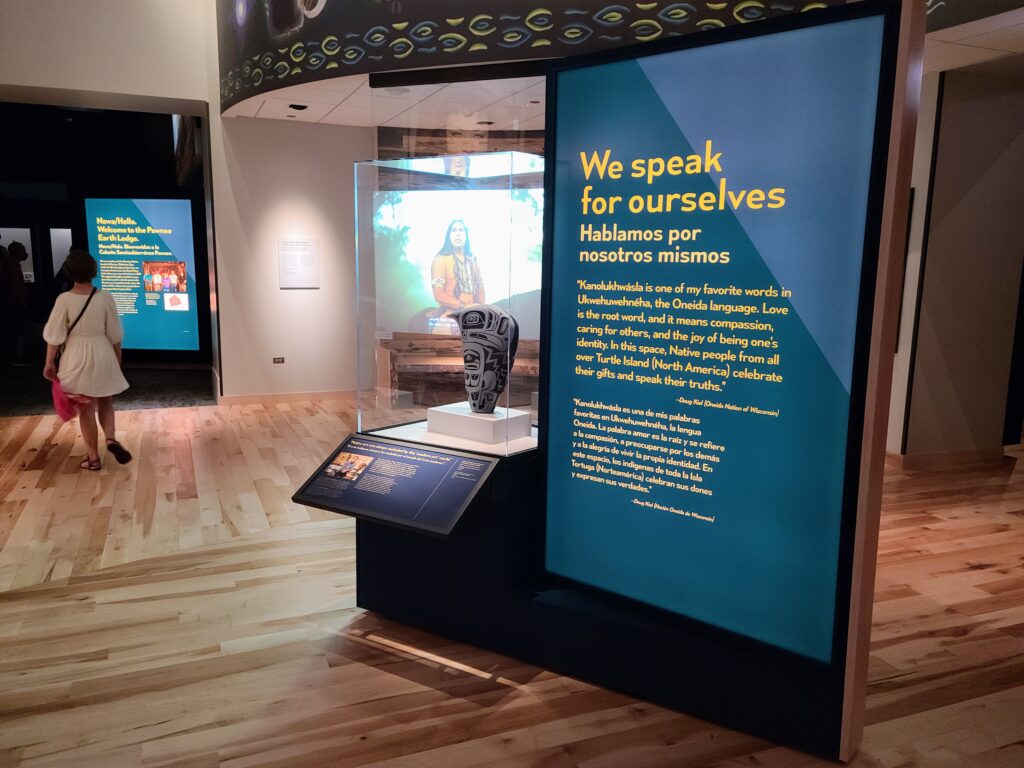 A photograph features a spacious museum exhibit. In the middle is a case that holds an object with white-and-black designs on it. A large blue wall on its right features text in English and Spanish that reads, “We speak for ourselves. ‘Kanolukhwásla is one of my favorite words in Ukwehuwehnéha, the Oneida language. Love is the root word, and it means compassion, caring for others, and the joy of being one’s identity. In this space, Native people from all over Turtle Island (North America) celebrate their gifts and speak their truths.’—Doug Kiel (Oneida Nation of Wisconsin).”