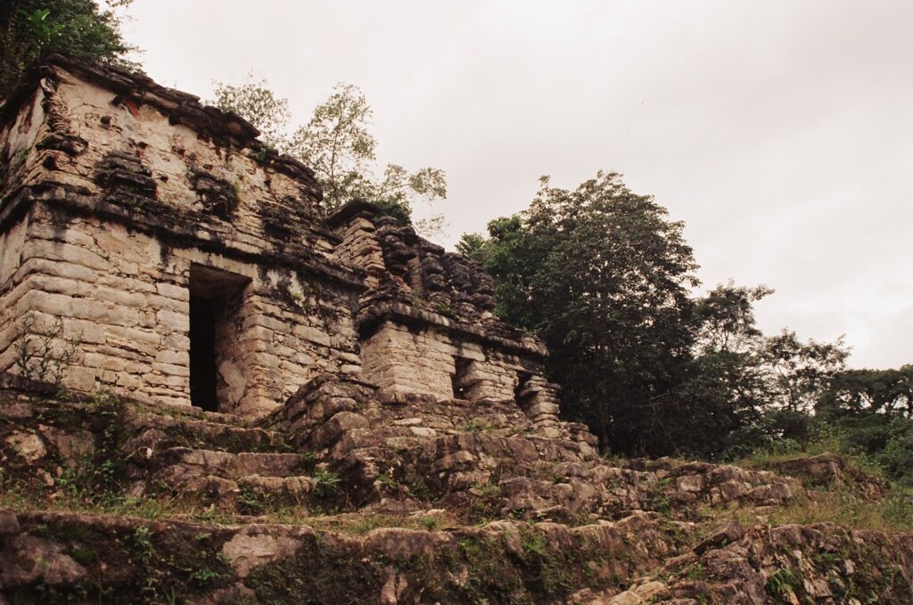 A partially ruined stone building stands on a rocky cliffside surrounded by trees.