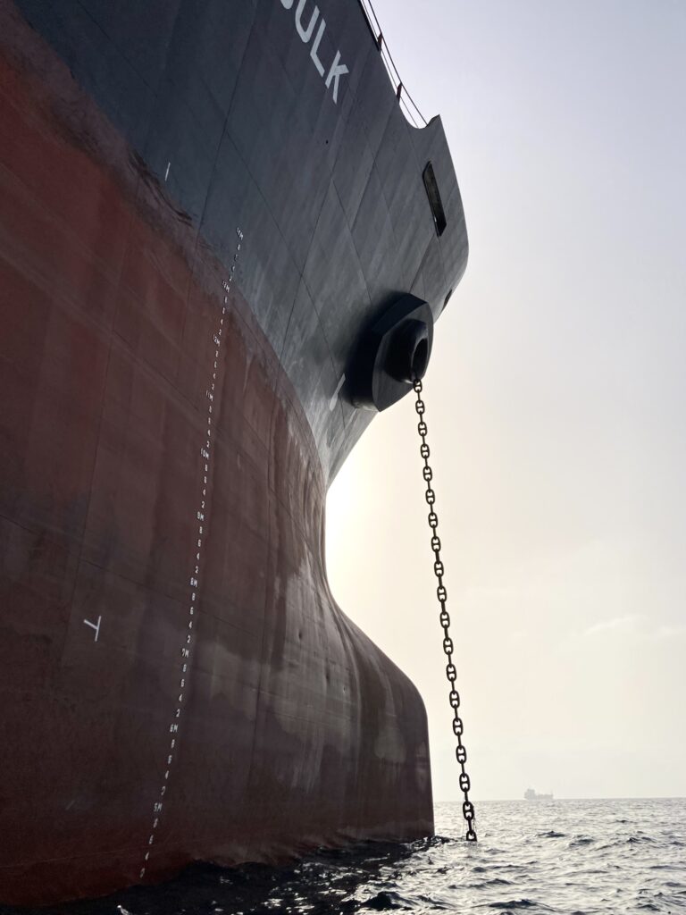 A photograph features the side of a large ship—blue on its top half and red on its bottom half—with a long chain descending from the side of the ship into dark blue water.