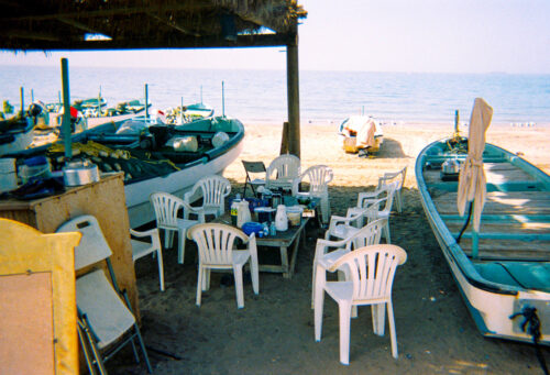 A photograph features a seaside cluster of small white and teal boats filled with miscellaneous objects parked on the sand around white plastic chairs. The chairs are arranged around a wooden table that holds jars, cups, and bowls.