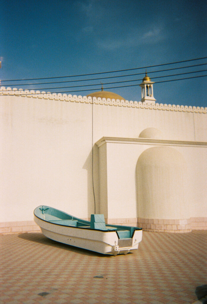 A photograph features a small white boat with a teal interior parked on a floor of gray and brown tile in front of a tall beige stone wall. A white spire topped with gold is visible behind the wall.