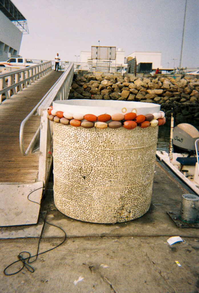 A photograph features a beige water tank with its top circumference wrapped with strings of orange cylindrical buoys. The tank sits at the base of a wooden slatted ramp that leads up to a boardwalk.