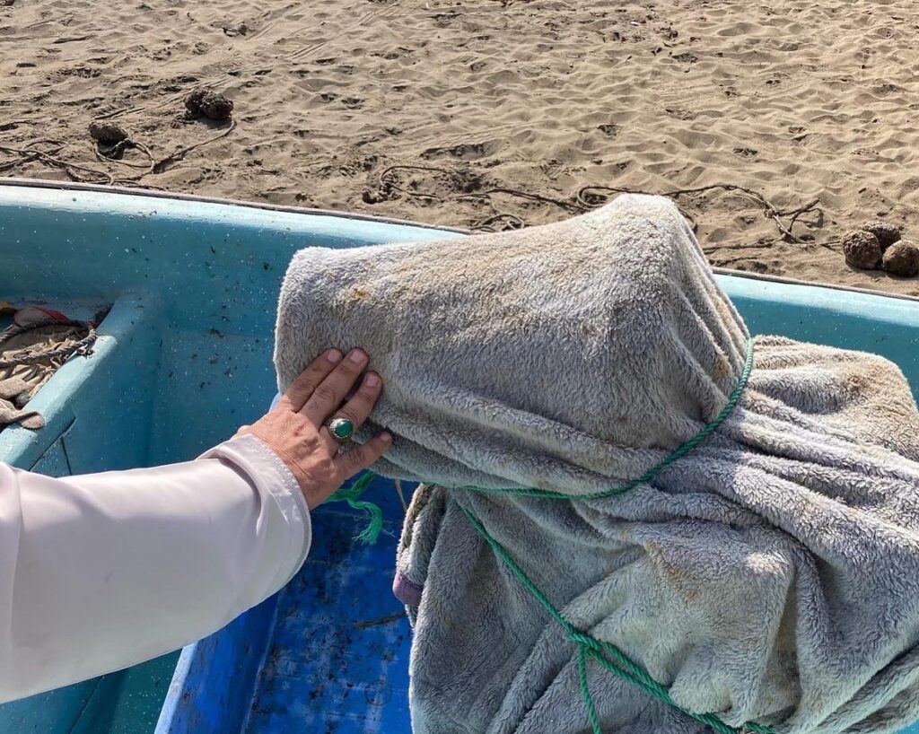 A photograph features a hand holding up an object wrapped in a dirty blanket and tied with green nylon rope. The object is sitting in a boat with a teal interior that has been pulled onto a sandy beach.