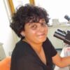 A photograph features a seated person with short curly black hair, pierced ears, and a nose ring wearing a black t-shirt. They look up from a microscope in front of them to smile at the viewer.
