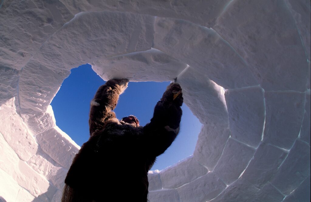 From below, a photograph features a person in a brown fur coat attaching a large ice block to an in-progress igloo from inside, with a blue sky above.