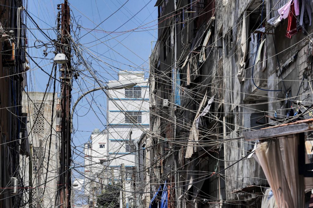 A photograph features a narrow street lined with tall white and beige buildings. Left of the photo’s center, a large telephone pole has numerous wires extending from it.