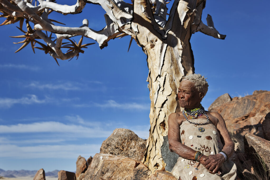 A photograph features an older person with short, kinky white hair looking to the right and leaning against a leafless tree. A rocky mountain crest and blue sky are in the background.