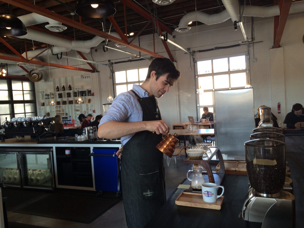 A photograph features a person wearing a collared shirt and apron in a large kitchen pouring coffee from a kettle into one of several white mugs on a wooden counter.