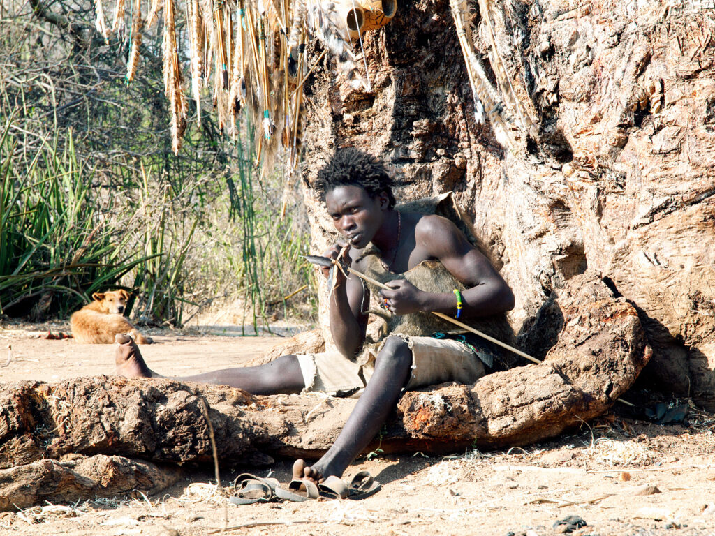 A photograph features a person sitting on an exposed tree root in front of a tree. They are holding a spear and looking into the distance.