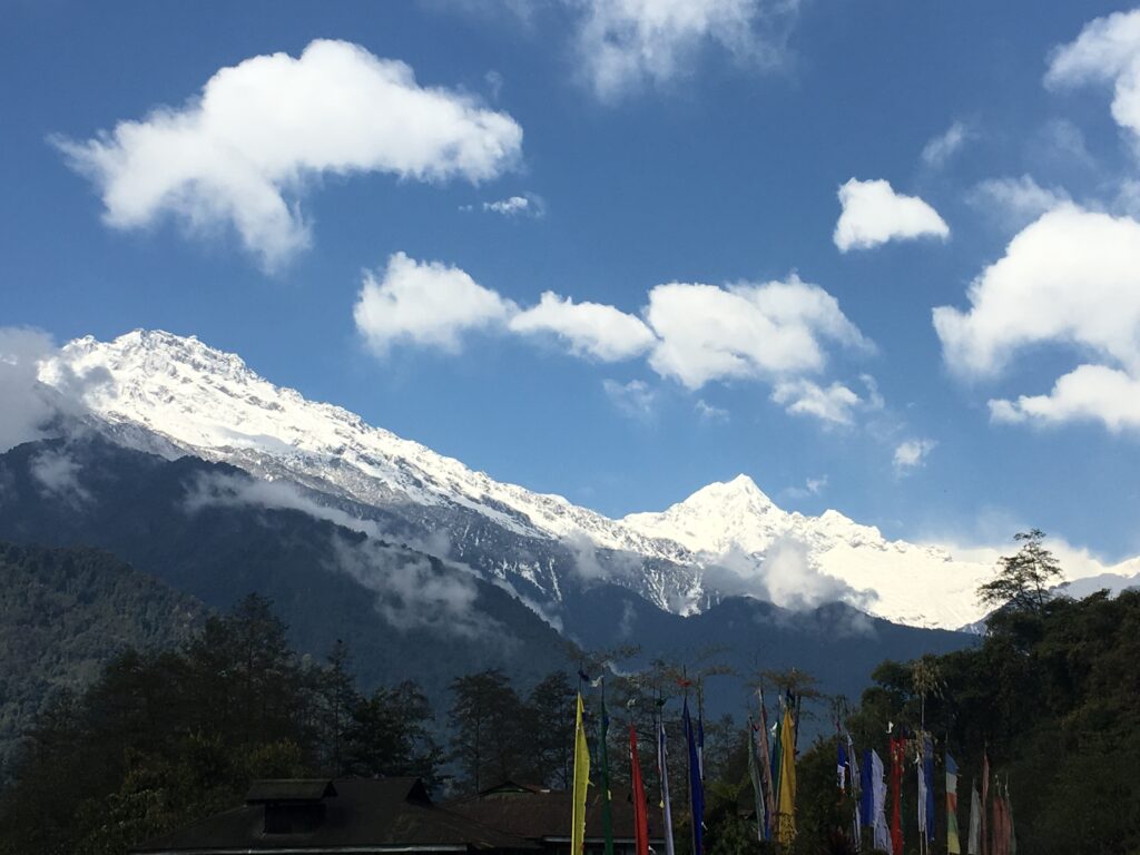 A zoomed-out photograph features white-capped mountains towering in the distance against a blue sky with light clouds. Flags of different colors and dark, sparse shrubbery are in the foreground.