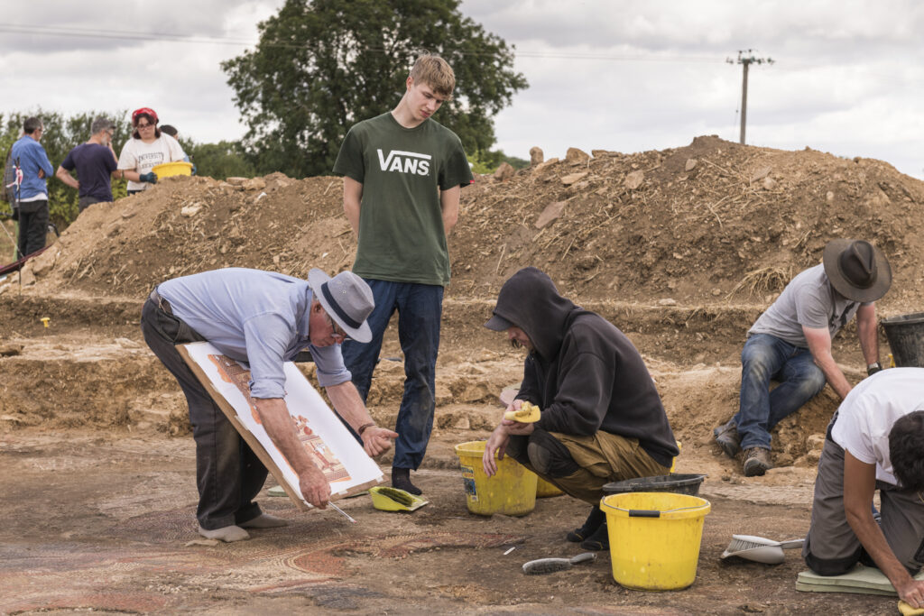 A photograph features, among other people, an older person with a brimmed hat showing an illustration to two younger people on an archaeological site.