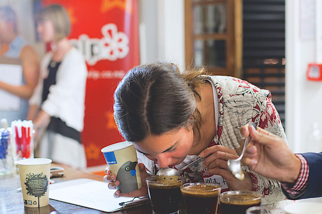A photograph shows a person wearing a cardigan bent over smelling a frothy dark liquid in a glass cup, to which they are holding a silver spoon.