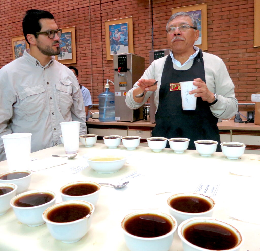 A photograph shows one person looking at another who holds a white cup in one hand and gestures with a spoon in the other. They stand at a table that holds more than a dozen white cups filled with dark liquid.