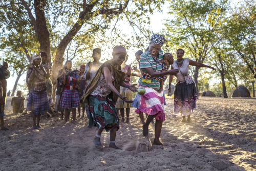 A photograph features a group of people in patterned clothing gathered on a sandy plot with domed huts visible in the distance. In front, two people dance as others cheer.