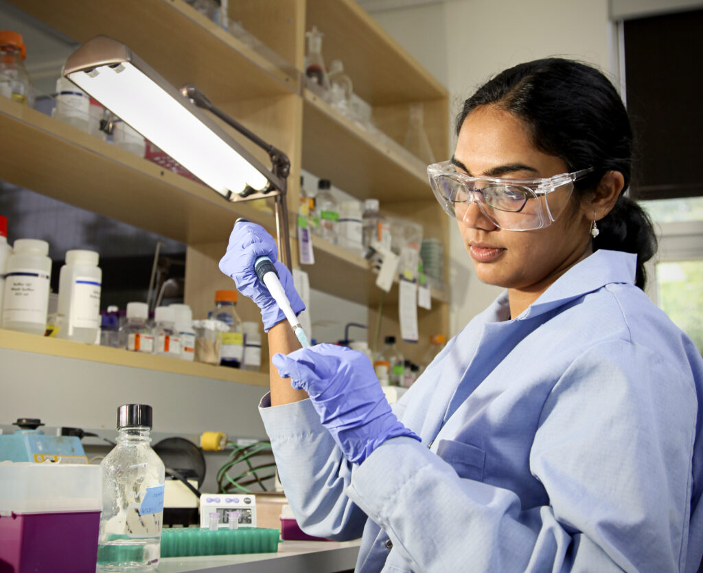 A photograph features a person with a long black ponytail wearing clear goggles and a blue lab coat and gloves. They are holding a syringe in a lab.
