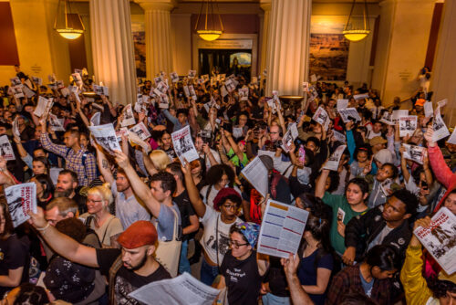 A photograph features a large crowd inside an atrium holding up flyers emblazoned with capitalized red text that reads “Decolonize This Museum.”