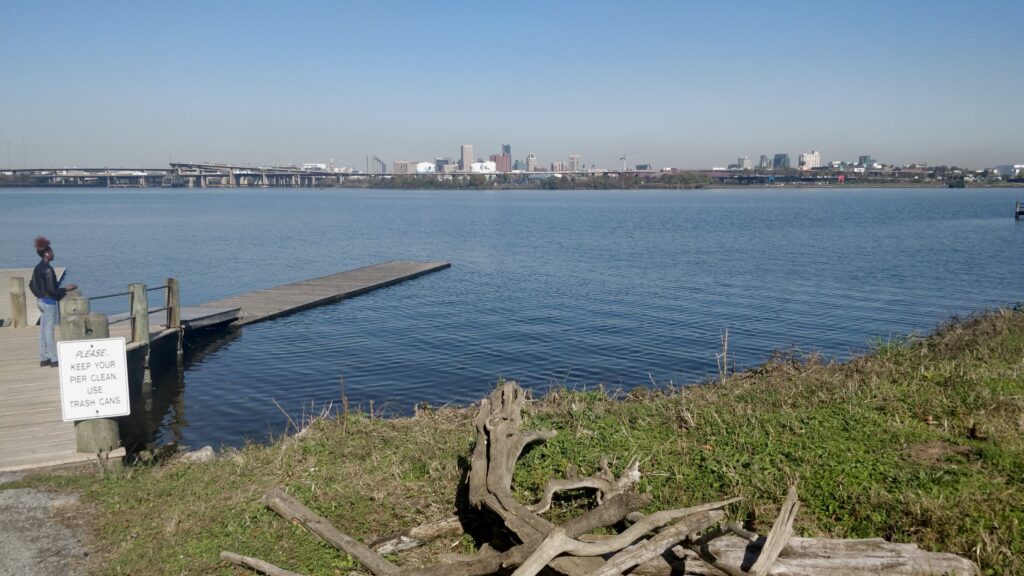 A photograph features a large body of water with a cityscape visible in the distance. On the left side, a person stands on a wooden pier near a white sign that reads: “PLEASE KEEP YOUR PIER CLEAN. USE TRASH CANS.”