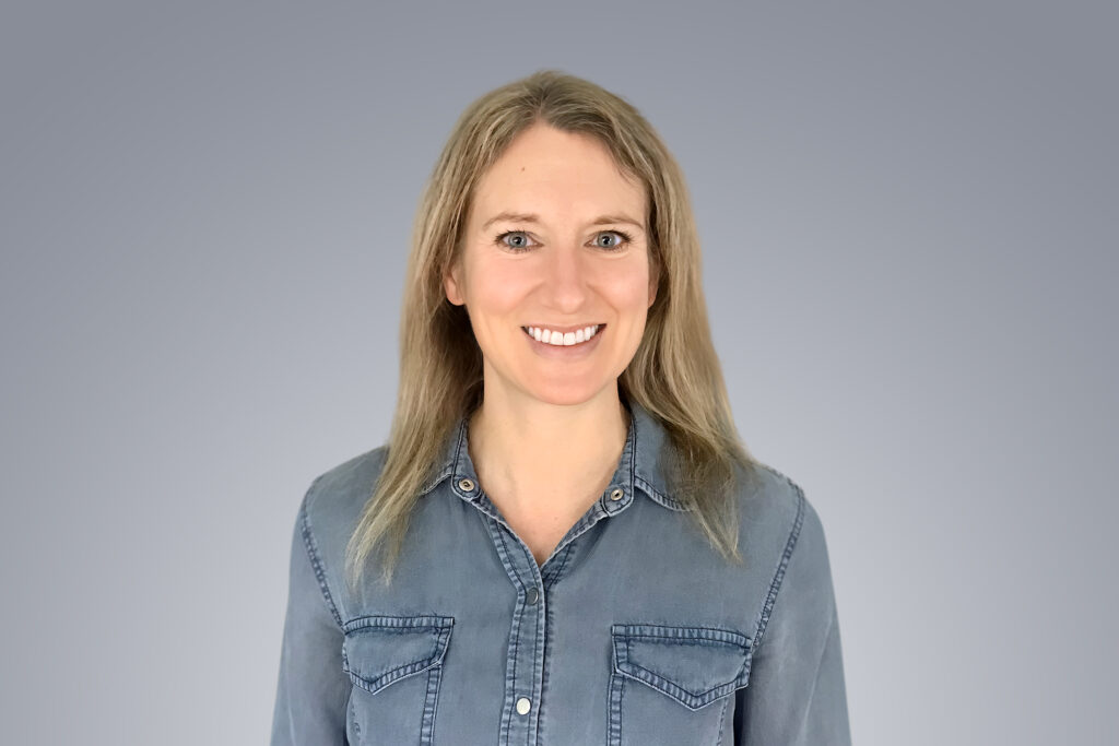 A White woman in a blue shirt smiles before the camera.