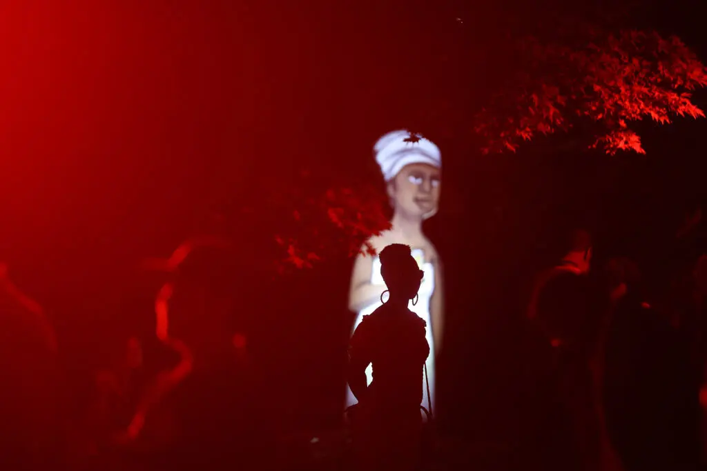 A photograph features a dark scene with red light shining from the left to silhouette a crowd of people under an overhanging tree. In the center, a person wearing hoop earrings and a head wrap poses in front of a projected illustration of a person in a white dress and head wrap.