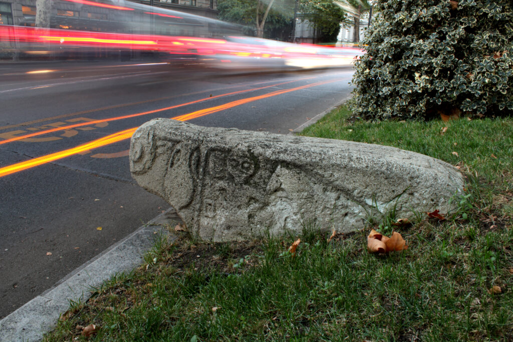 A large, sheep-shaped stone with etchings on its side sits on grass alongside a road where a car speeds by in a blur of orange lights.