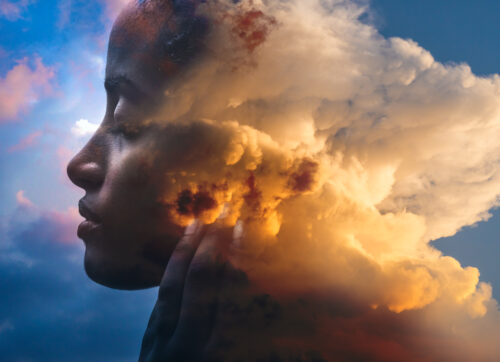 A picture features a sky with a large, slightly orange, billowing cloud at its center that morphs on its left side to look like the side profile of dark-skinned woman’s face. To the left of her is a bright-blue sky with clouds.