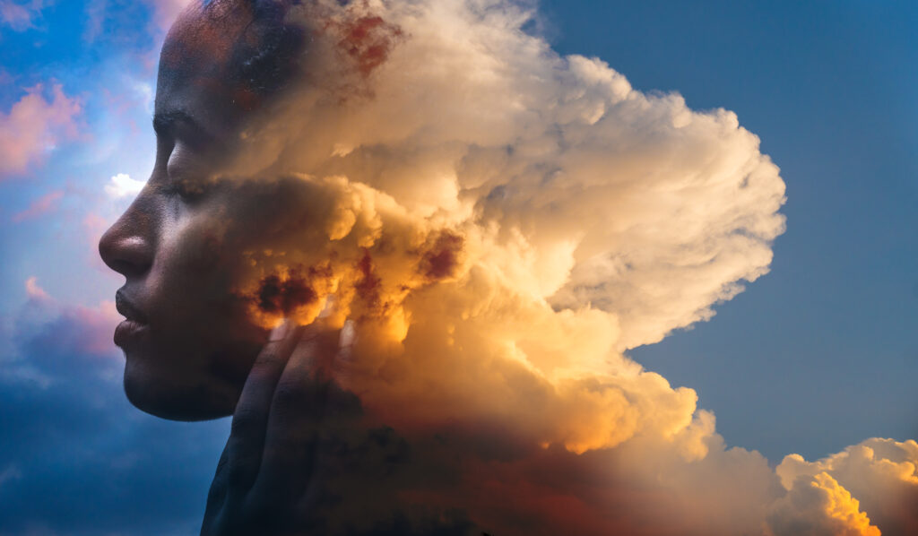 A picture features a sky with a large, slightly orange, billowing cloud at its center that morphs on its left side to look like the side profile of dark-skinned woman’s face. To the left of her is a bright-blue sky with clouds.