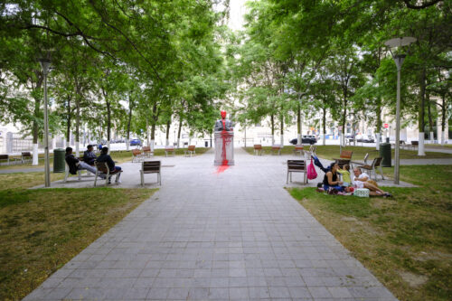 A photograph features a square area of concrete tiling in a park’s center with bench seating around its perimeter. At the center of the square is a stone statue of a man’s bust covered in red paint.