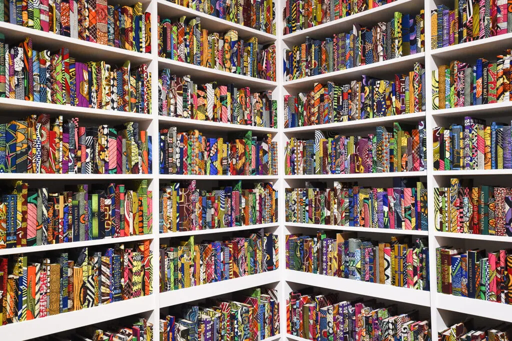 A photograph features the corner of a large bookcase filled with books. The books’ various multicolored covers evoke patterns common on African wax print fabrics.