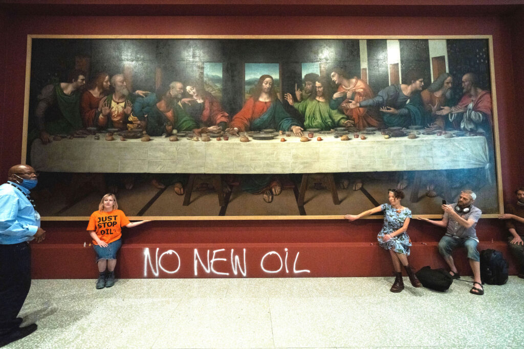 Three people sit in a museum with their hands on the frame of a large painting depicting people sitting at a long table. Under the painting, the words “No New Oil” are spray-painted on the wall.