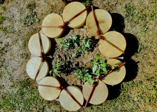 The photograph features an aerial shot of a circle of drums on dirt ground sparingly covered with grass and green leaves. Wooden drum sticks are arranged across the drums’ tops to form a star shape.