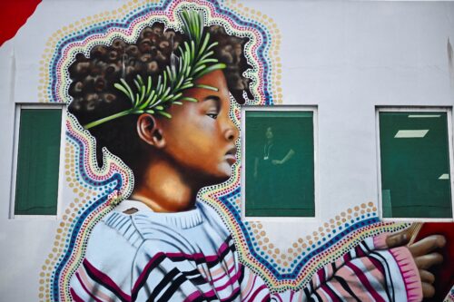A photograph features a building’s white wall covered with a large colorful mural of a curly haired young person with a green sprig tucked behind their ear. The mural surrounds a window through which a woman can be seen standing with her hand on her hip.