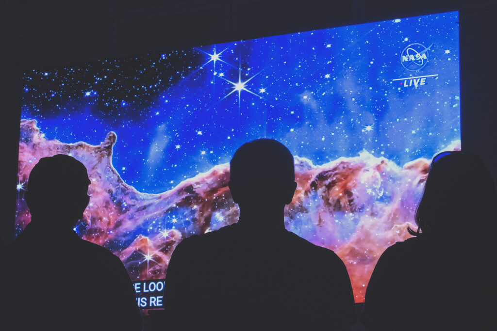A photograph shows the silhouettes of three people against a large screen with an image of a colorful galaxy across it.