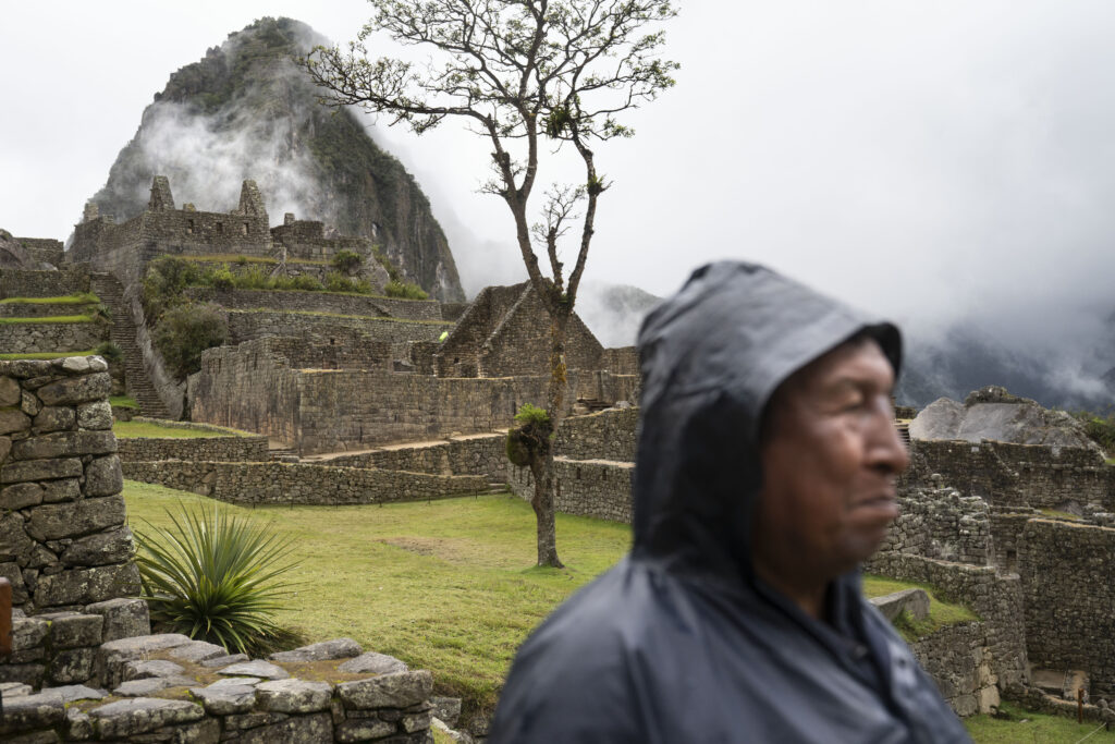 A photograph features a person in a hooded windbreaker in the foreground looking left, away from a large tiered moss-covered stone structure in the background.